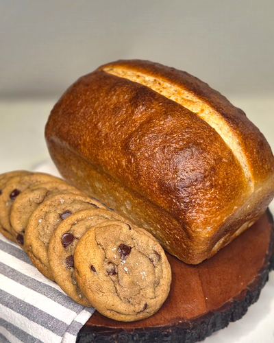 Loaf of bread and chocolate chip cookies on a wooden board with a striped towel.